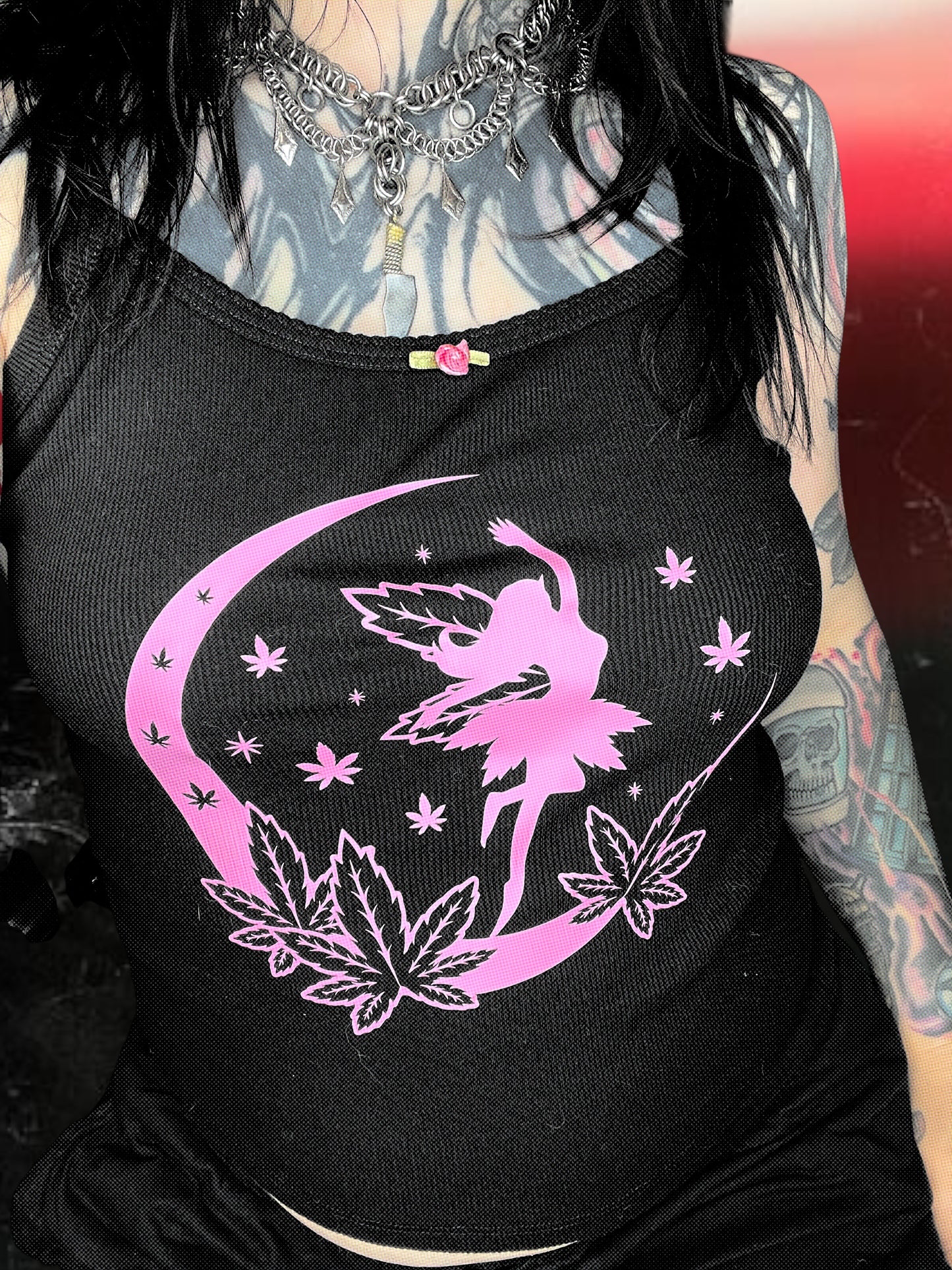 A person wearing a black tank top with a pink fairy design and a crescent moon, along with some leafy plants.