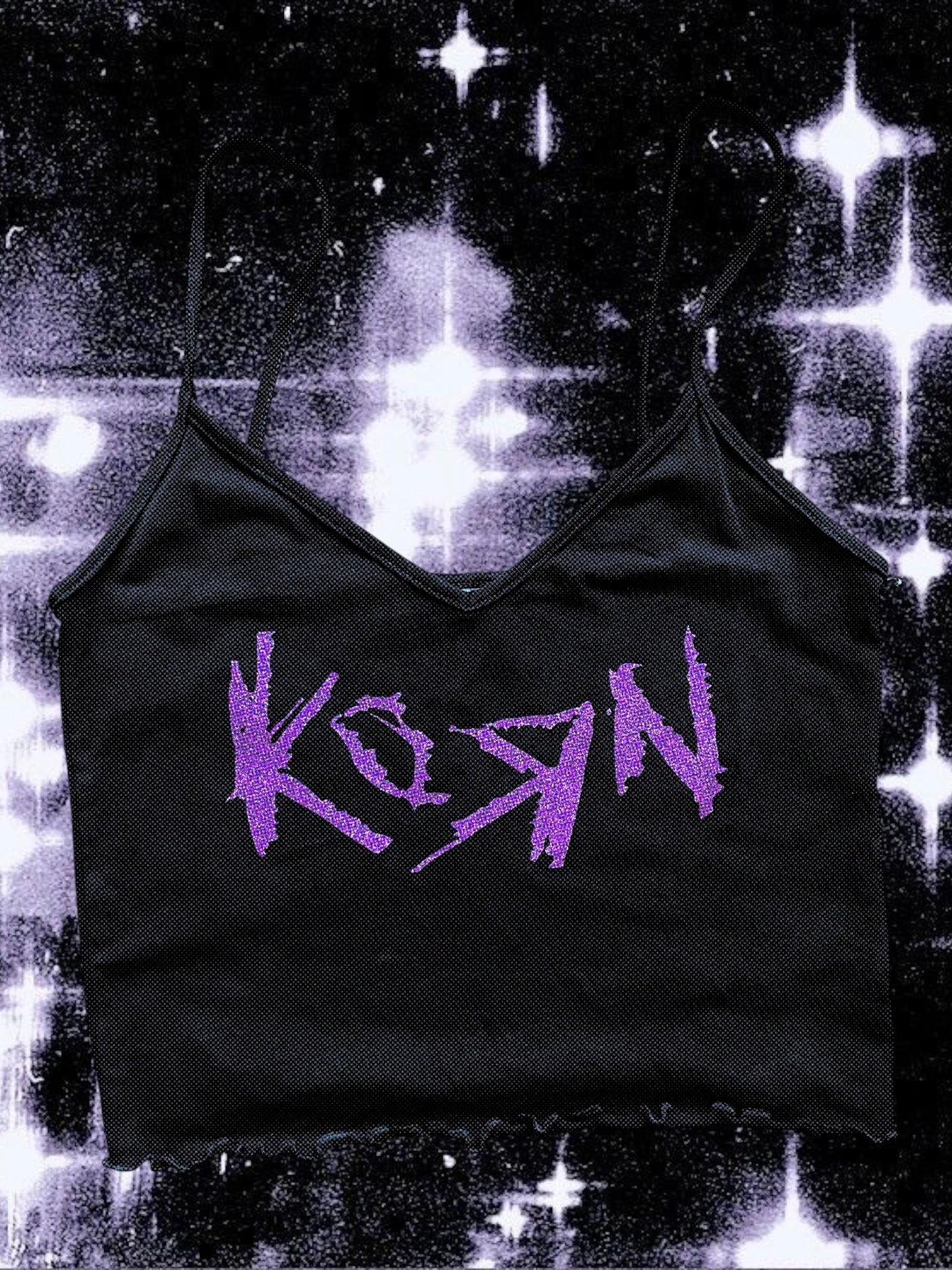 Black cropped spaghetti strap cami with a raw hem, featuring the Korn band logo printed in glittery purple. The shirt is laid flat against a cosmic black-and-white starry background.