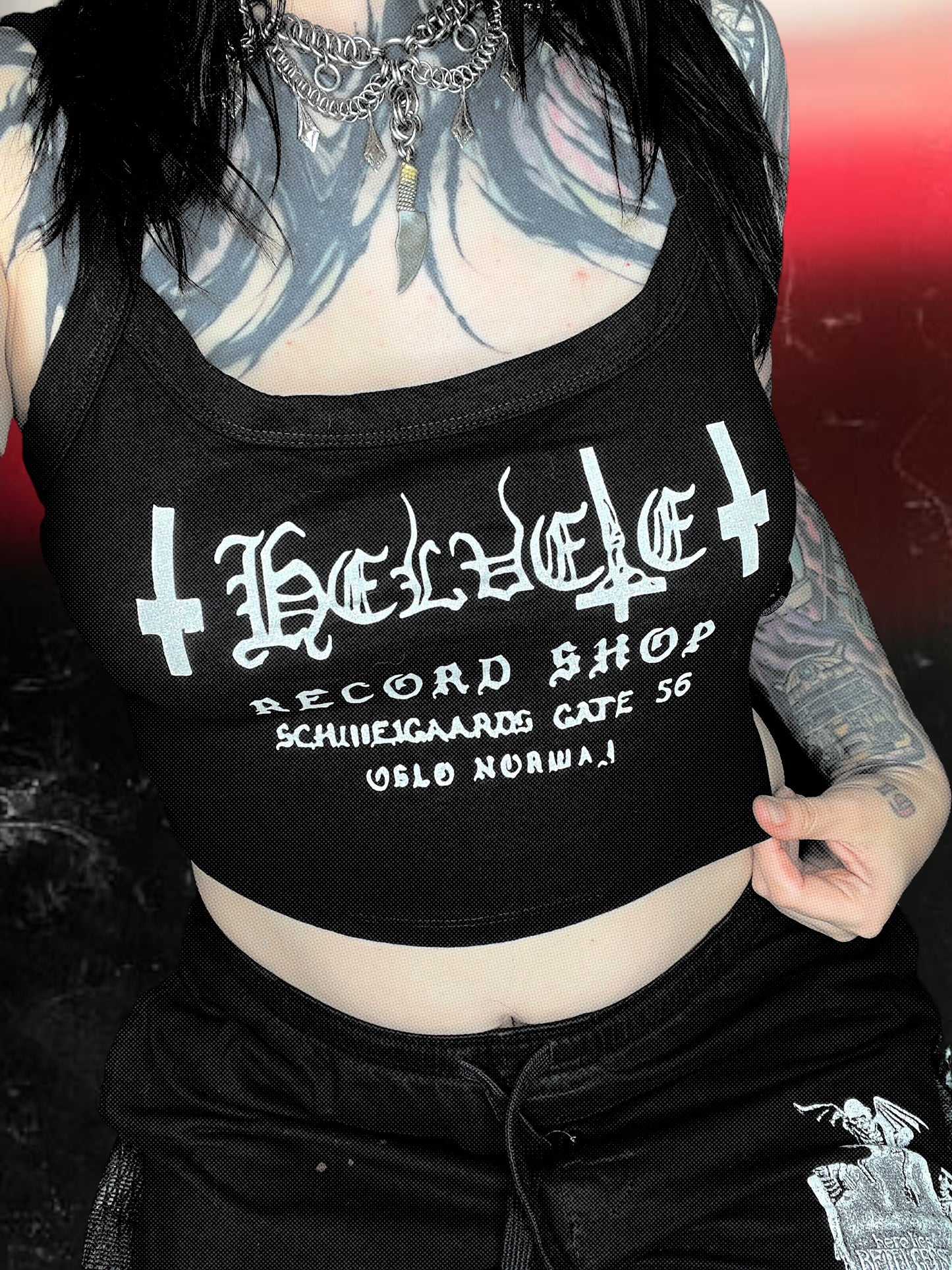 Black cropped spaghetti strap cami with “Helvete Record Shop” printed in white glitter vinyl. The design includes gothic text, upside-down crosses, and the address "Schweigaards Gate 56, Oslo Norway." Shirt is laid flat on a cosmic black-and-white starry background.