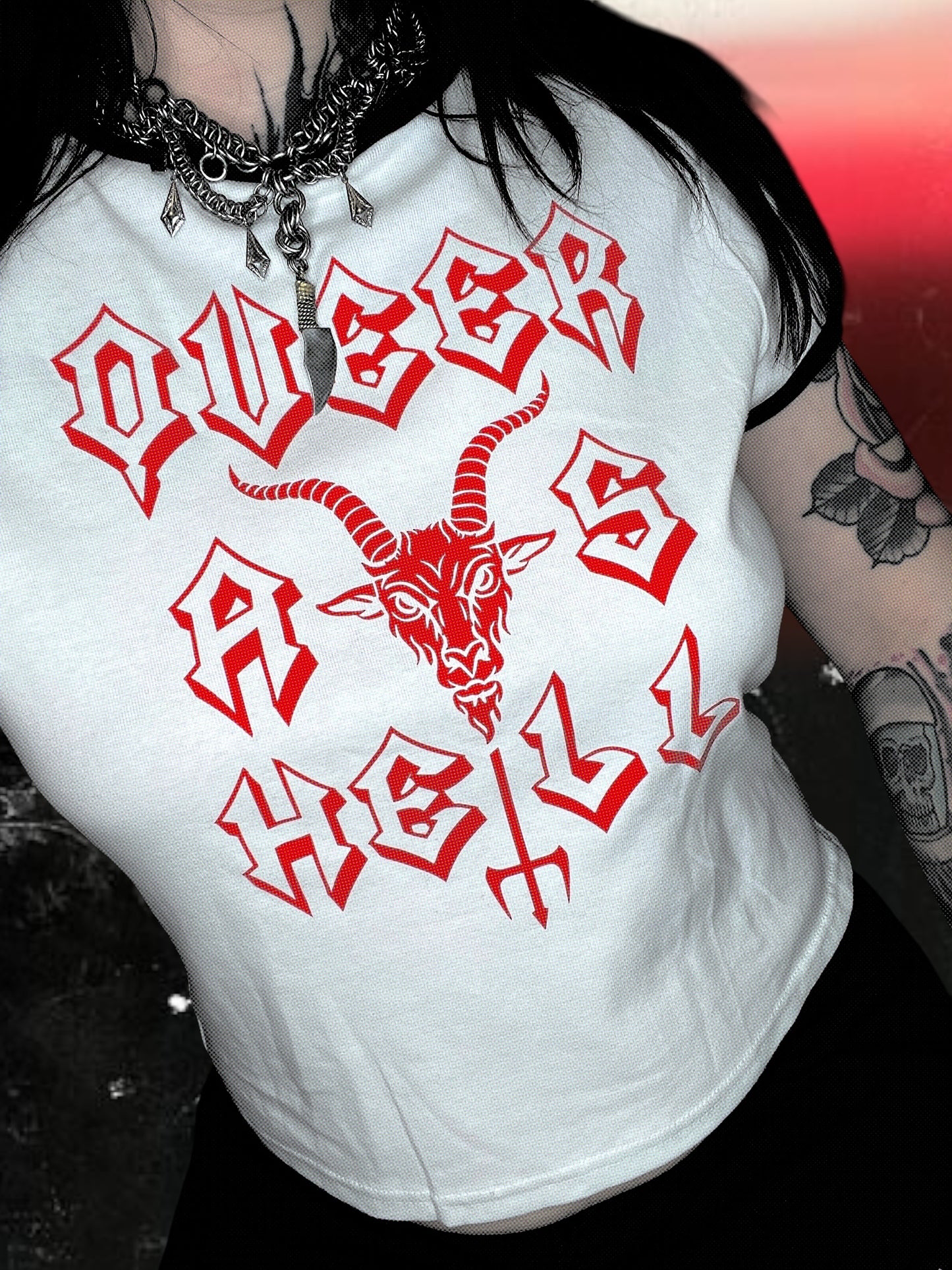 White baby tee with black ringer trim and red gothic-style text that says “QUEER AS HELL.” The design includes a goat head above a trident that forms part of the word “HELL.” The shirt is laid flat on a cosmic black-and-white starry background; lighting makes the shirt appear slightly blue.