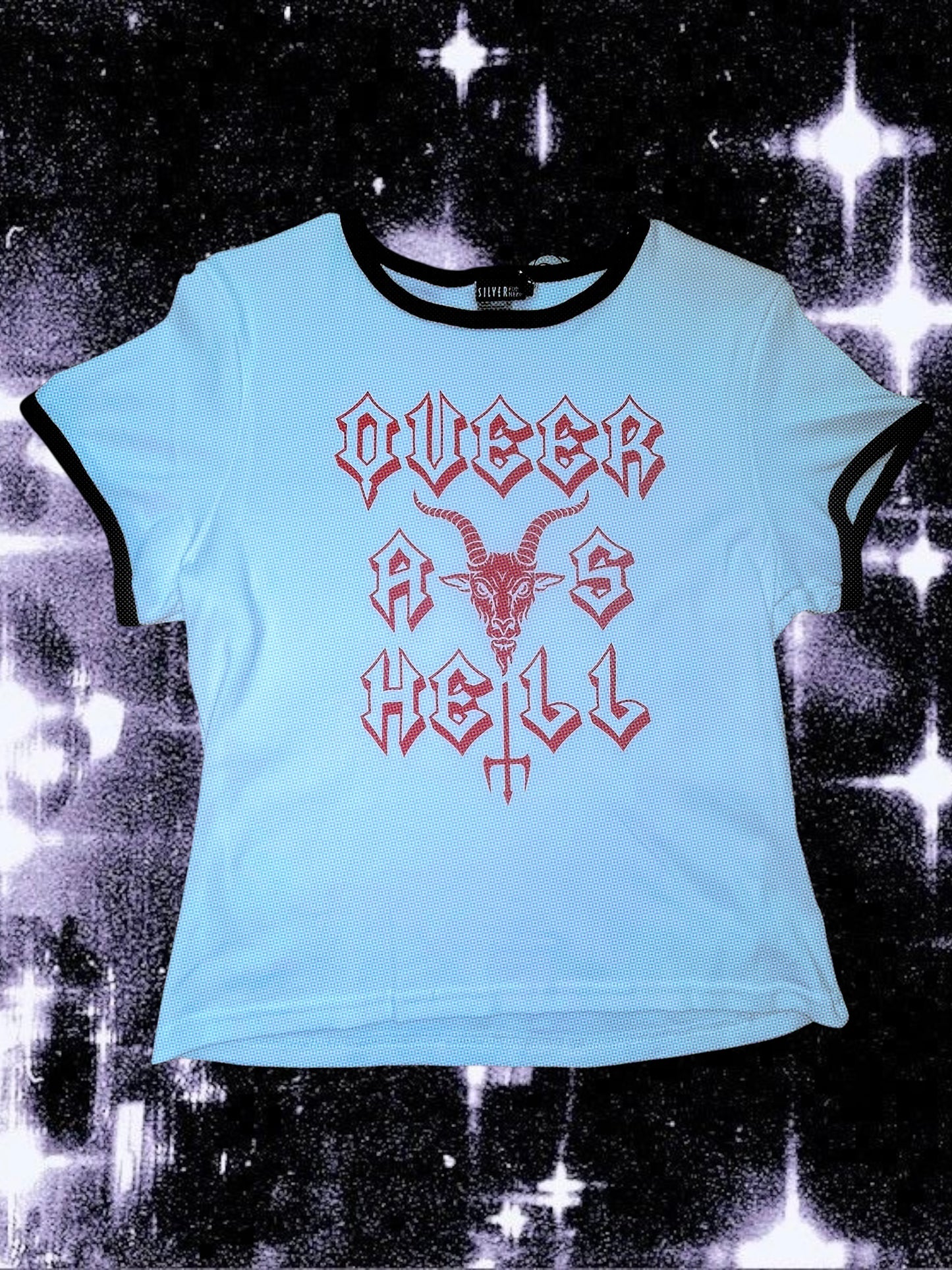White baby tee with black ringer trim and red gothic-style text that says “QUEER AS HELL.” The design includes a goat head above a trident that forms part of the word “HELL.” The shirt is laid flat on a cosmic black-and-white starry background; lighting makes the shirt appear slightly blue.