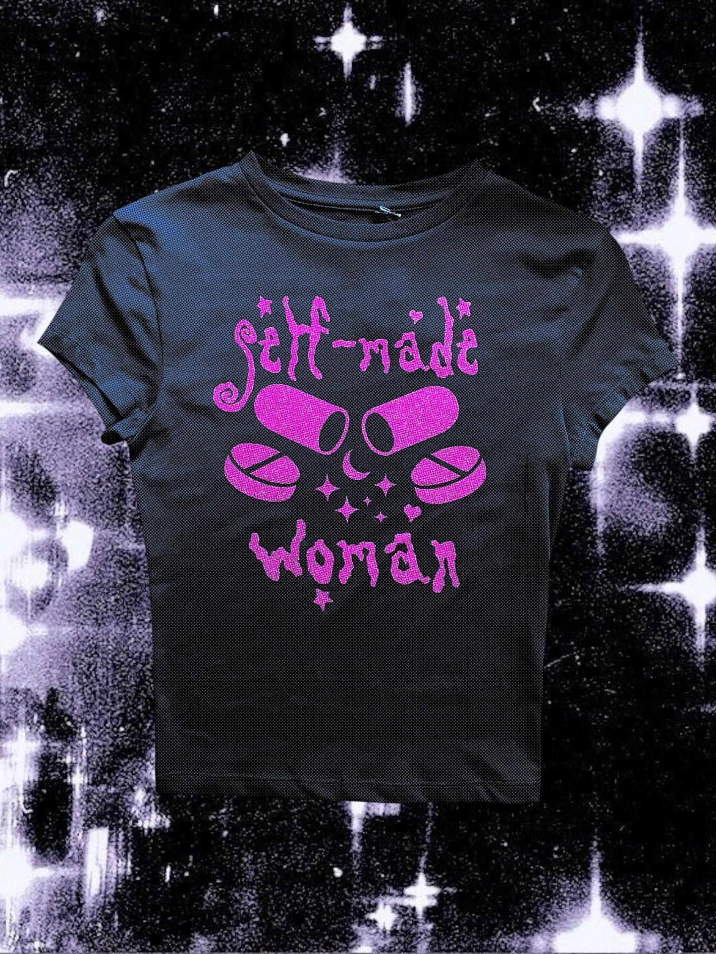 Black fitted baby tee with pink glitter vinyl print showing two capsules crossed like bones, surrounded by sparkles. The text “self-made woman” is written above and below the pills in a gothic, whimsical font. The shirt is laid flat against a cosmic black-and-white starry background.