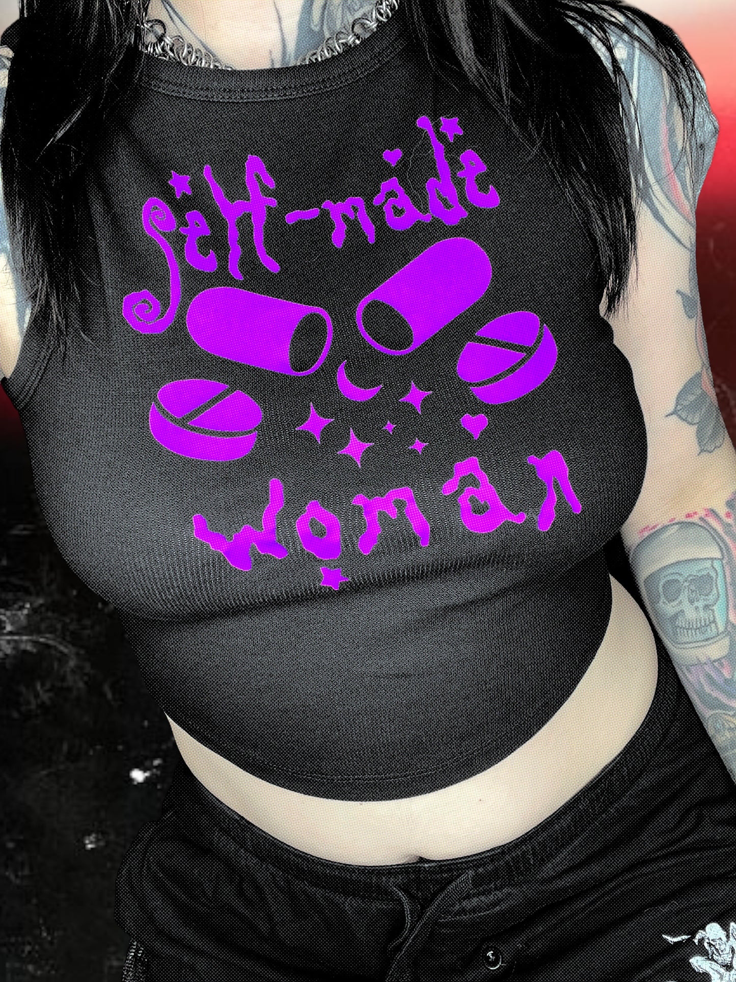 Black cropped tank top with a purple vinyl design that says “self-made woman” in a witchy gothic font. The center graphic shows two large capsule pills crossed, surrounded by sparkles and smaller round tablets. Shirt is laid flat over a black-and-white starry cosmic background.