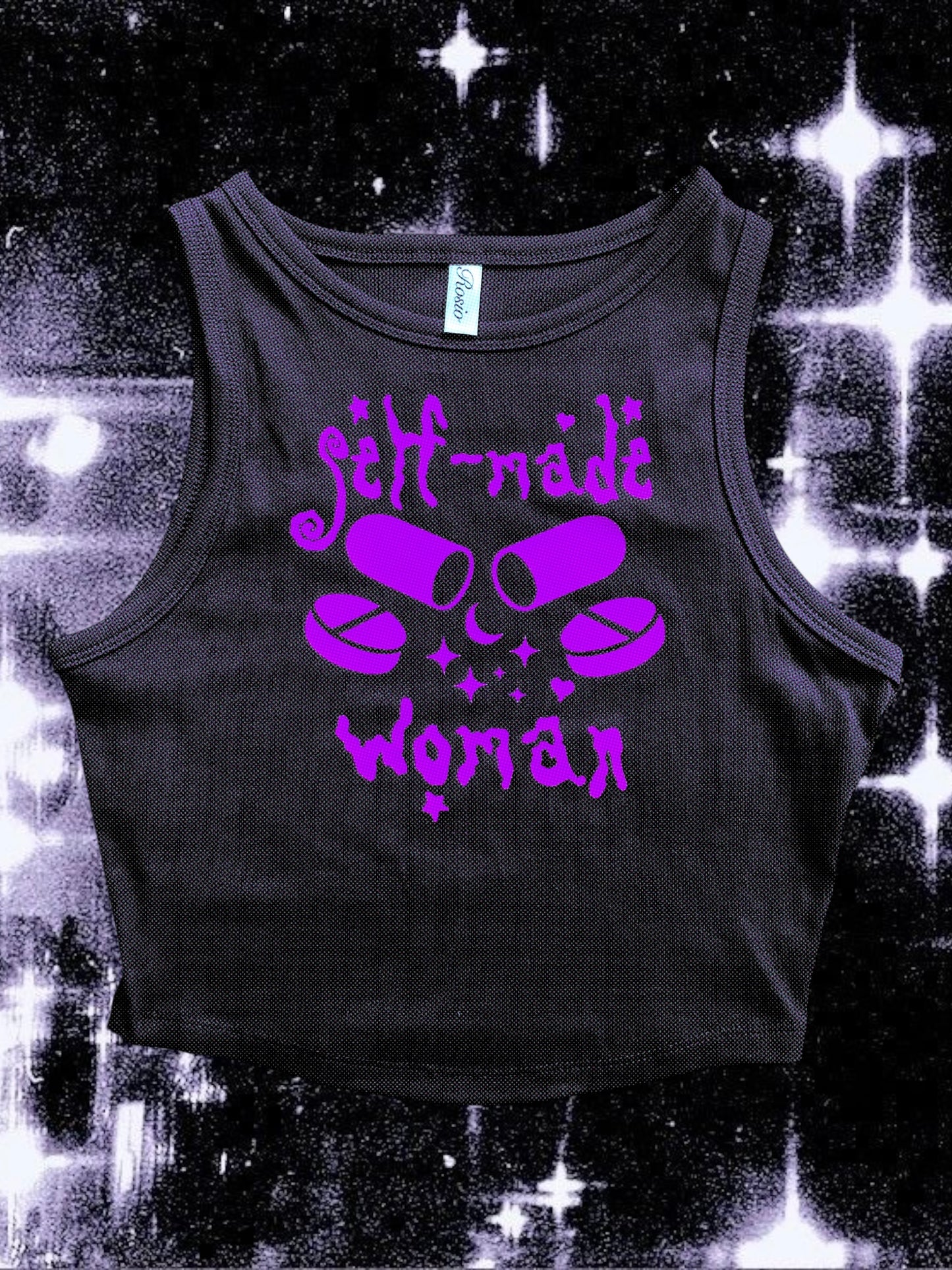 Black cropped tank top with a purple vinyl design that says “self-made woman” in a witchy gothic font. The center graphic shows two large capsule pills crossed, surrounded by sparkles and smaller round tablets. Shirt is laid flat over a black-and-white starry cosmic background.