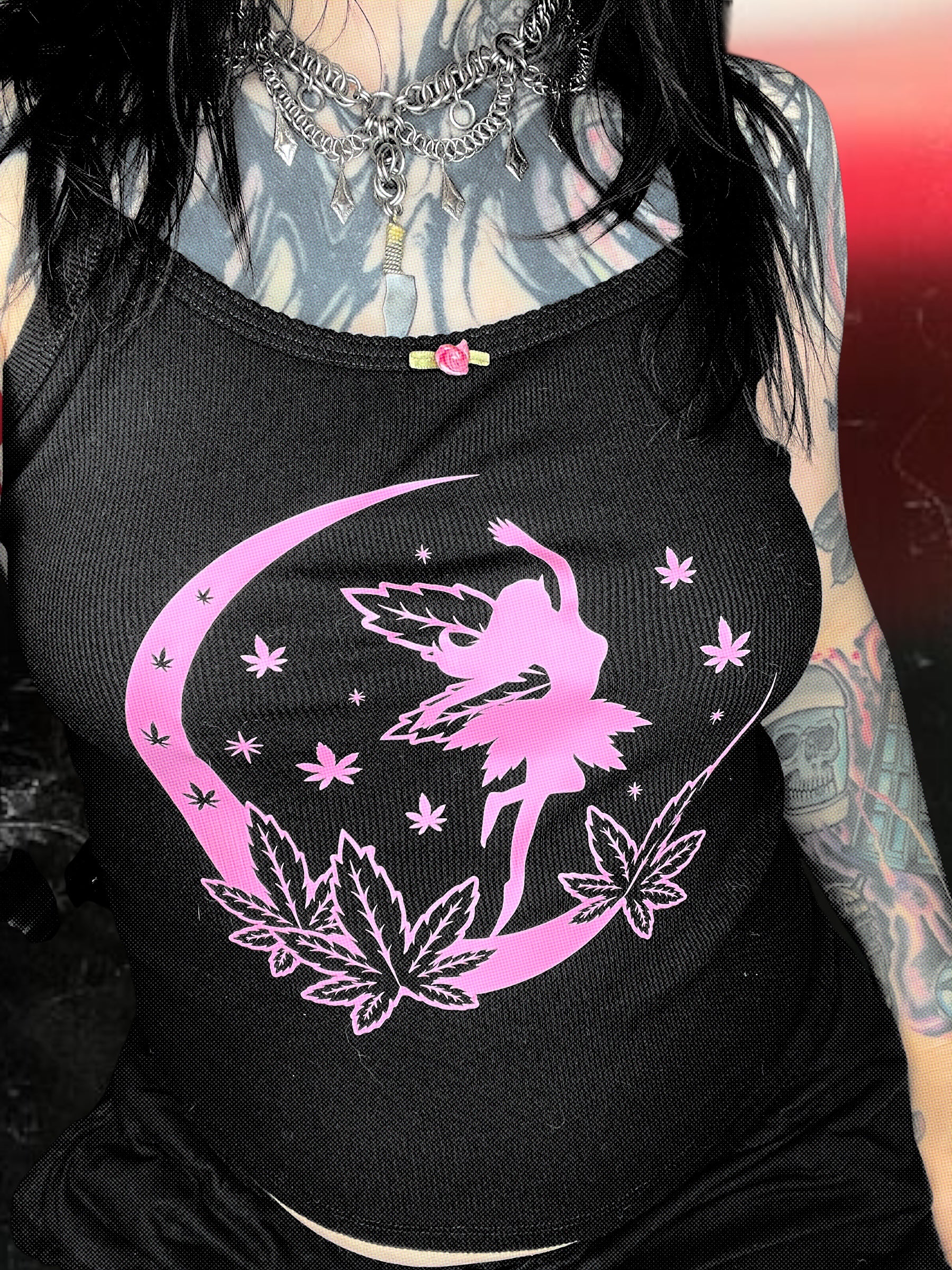 A person wearing a black tank top with a pink fairy design and a crescent moon, along with some leafy plants.
