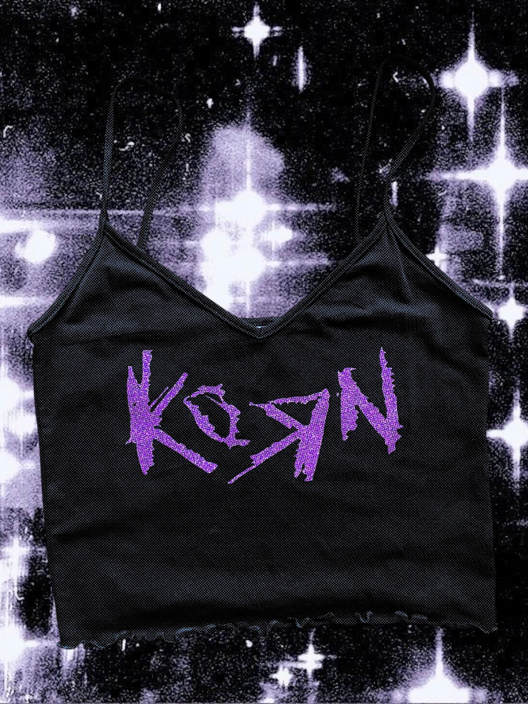Black cropped spaghetti strap cami with a raw hem, featuring the Korn band logo printed in glittery purple. The shirt is laid flat against a cosmic black-and-white starry background.