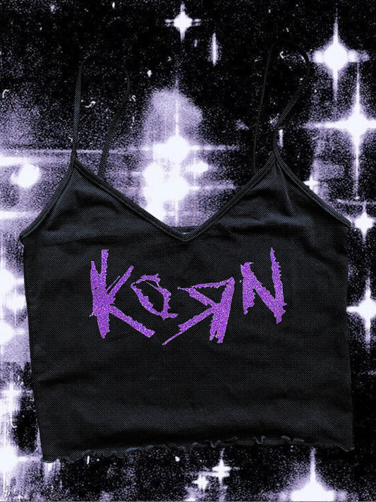 Black cropped spaghetti strap cami with a raw hem, featuring the Korn band logo printed in glittery purple. The shirt is laid flat against a cosmic black-and-white starry background.