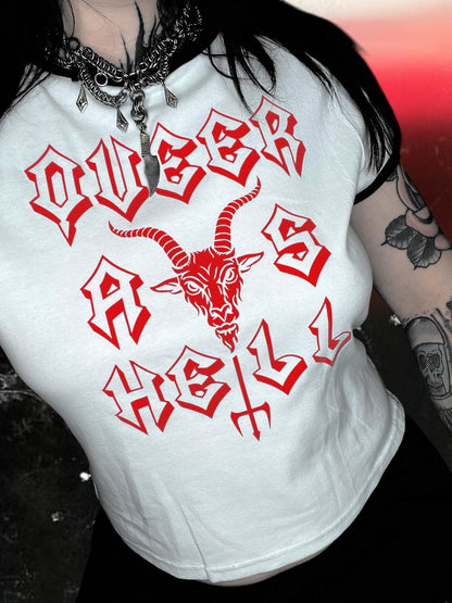 White baby tee with black ringer trim and red gothic-style text that says “QUEER AS HELL.” The design includes a goat head above a trident that forms part of the word “HELL.” The shirt is laid flat on a cosmic black-and-white starry background; lighting makes the shirt appear slightly blue.