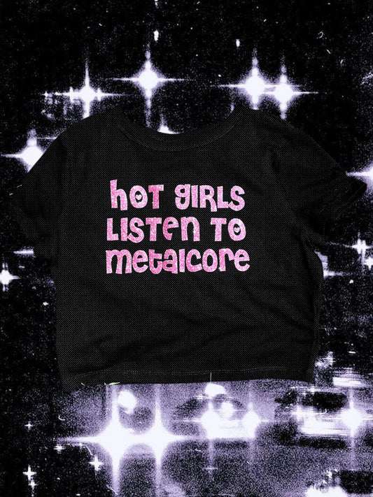Black baby tee with sparkly pink glitter text that says “hot girls listen to metalcore” in a chunky retro font. The shirt is laid flat against a cosmic, starry black-and-white background.