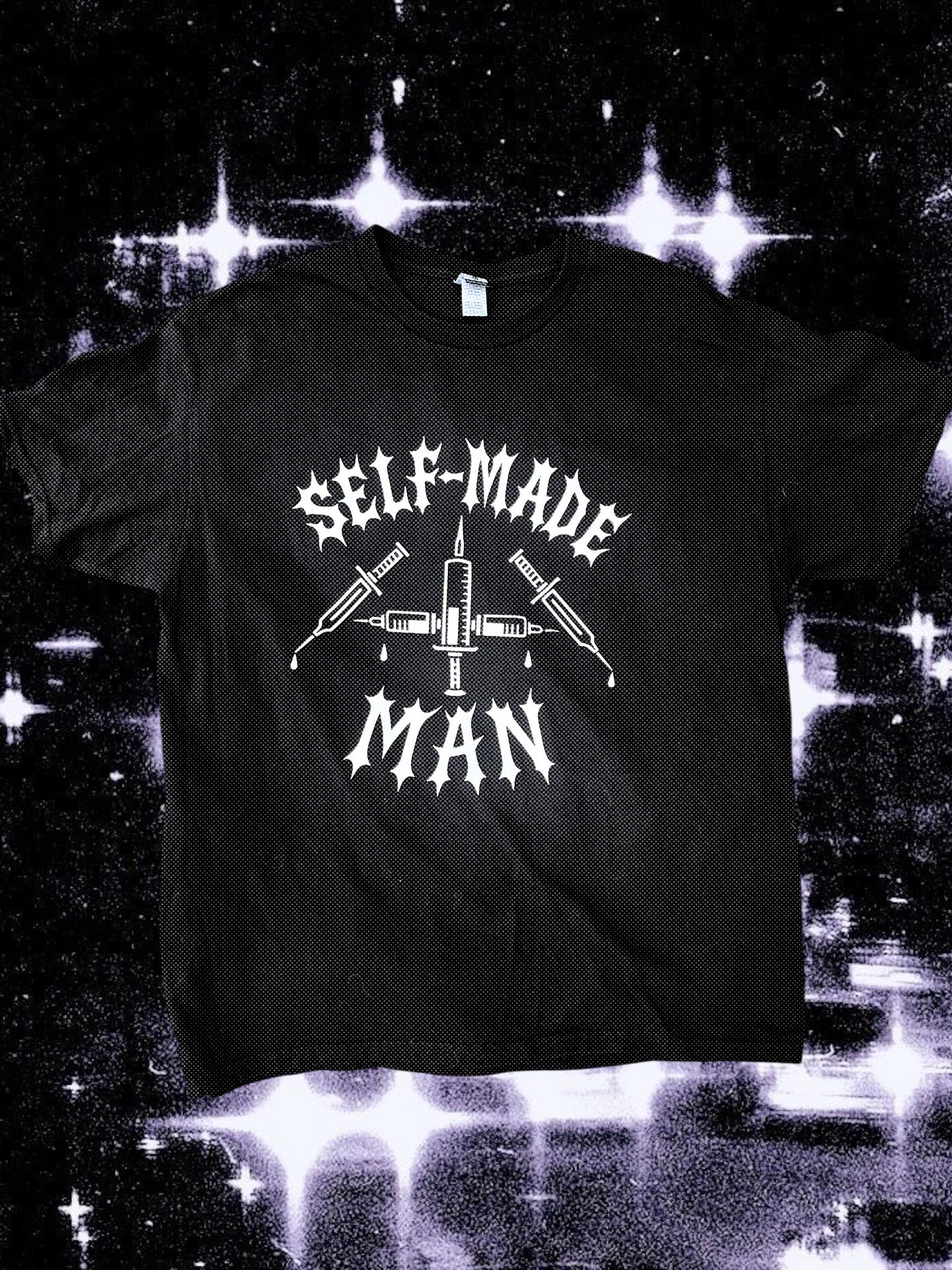 Black unisex short-sleeve t-shirt with white gothic-style text reading “SELF-MADE MAN.” The design includes four syringes arranged in an X shape with blood droplets. Shirt is laid flat on a black-and-white starry cosmic background.