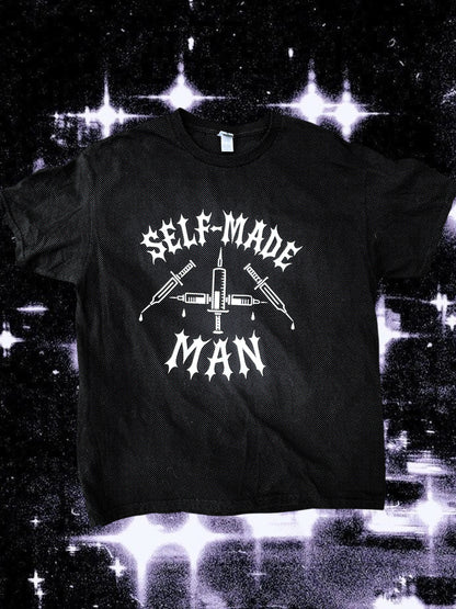 Black unisex short-sleeve t-shirt with white gothic-style text reading “SELF-MADE MAN.” The design includes four syringes arranged in an X shape with blood droplets. Shirt is laid flat on a black-and-white starry cosmic background.