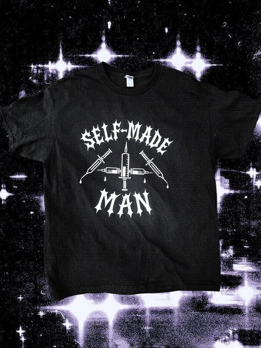 Black unisex short-sleeve t-shirt with white gothic-style text reading “SELF-MADE MAN.” The design includes four syringes arranged in an X shape with blood droplets. Shirt is laid flat on a black-and-white starry cosmic background.