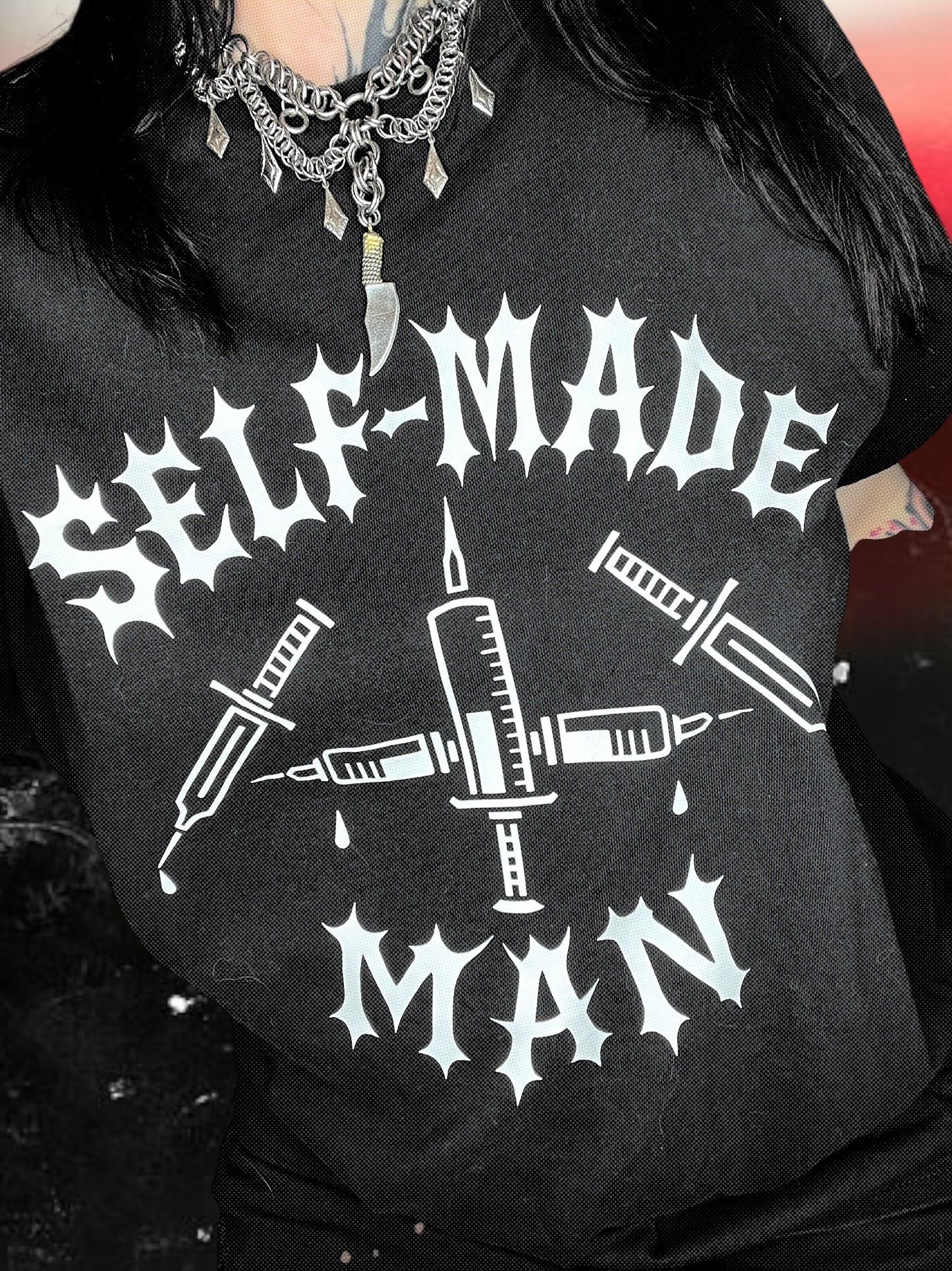 Black unisex short-sleeve t-shirt with white gothic-style text reading “SELF-MADE MAN.” The design includes four syringes arranged in an X shape with blood droplets. Shirt is laid flat on a black-and-white starry cosmic background.