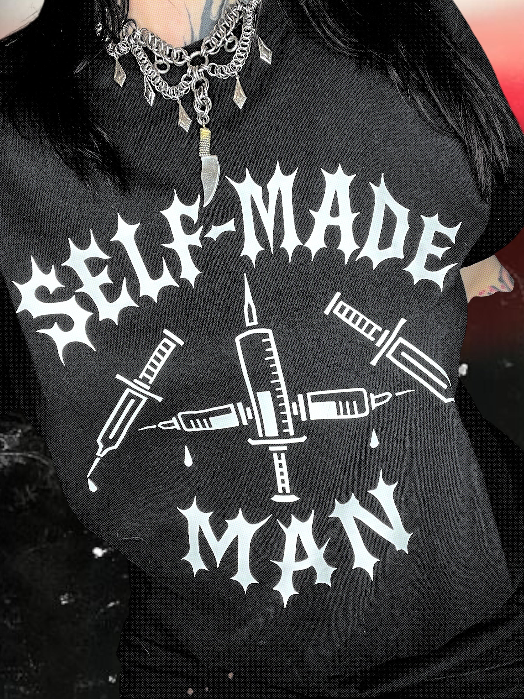 Black unisex short-sleeve t-shirt with white gothic-style text reading “SELF-MADE MAN.” The design includes four syringes arranged in an X shape with blood droplets. Shirt is laid flat on a black-and-white starry cosmic background.