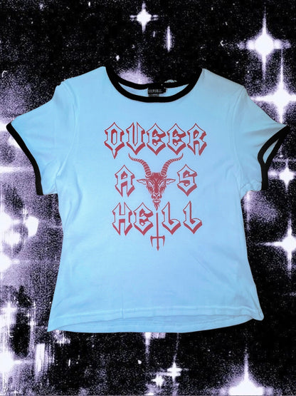 White baby tee with black ringer trim and red gothic-style text that says “QUEER AS HELL.” The design includes a goat head above a trident that forms part of the word “HELL.” The shirt is laid flat on a cosmic black-and-white starry background; lighting makes the shirt appear slightly blue.