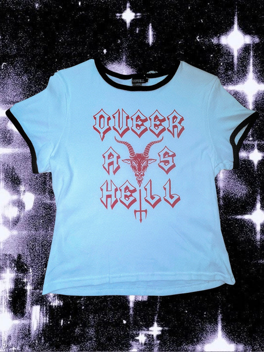 White baby tee with black ringer trim and red gothic-style text that says “QUEER AS HELL.” The design includes a goat head above a trident that forms part of the word “HELL.” The shirt is laid flat on a cosmic black-and-white starry background; lighting makes the shirt appear slightly blue.