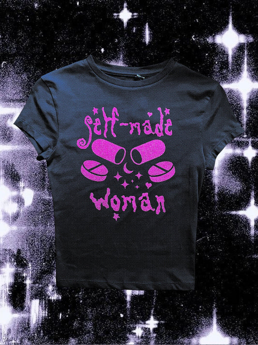Black fitted baby tee with pink glitter vinyl print showing two capsules crossed like bones, surrounded by sparkles. The text “self-made woman” is written above and below the pills in a gothic, whimsical font. The shirt is laid flat against a cosmic black-and-white starry background.