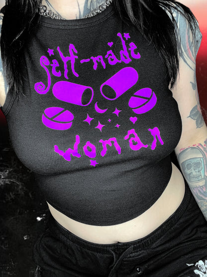 Black cropped tank top with a purple vinyl design that says “self-made woman” in a witchy gothic font. The center graphic shows two large capsule pills crossed, surrounded by sparkles and smaller round tablets. Shirt is laid flat over a black-and-white starry cosmic background.