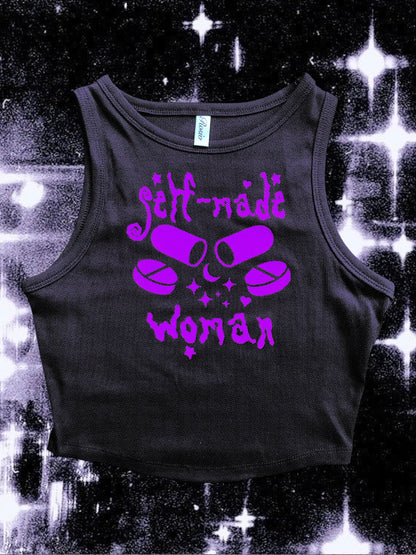 Black cropped tank top with a purple vinyl design that says “self-made woman” in a witchy gothic font. The center graphic shows two large capsule pills crossed, surrounded by sparkles and smaller round tablets. Shirt is laid flat over a black-and-white starry cosmic background.