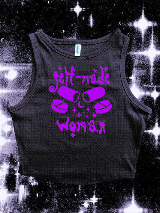 Black cropped tank top with a purple vinyl design that says “self-made woman” in a witchy gothic font. The center graphic shows two large capsule pills crossed, surrounded by sparkles and smaller round tablets. Shirt is laid flat over a black-and-white starry cosmic background.