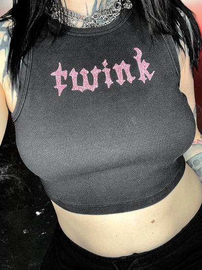 Black ribbed cropped tank top with pink glitter gothic text that reads “twink” across the chest. Shirt is laid flat against a black-and-white cosmic starry background.