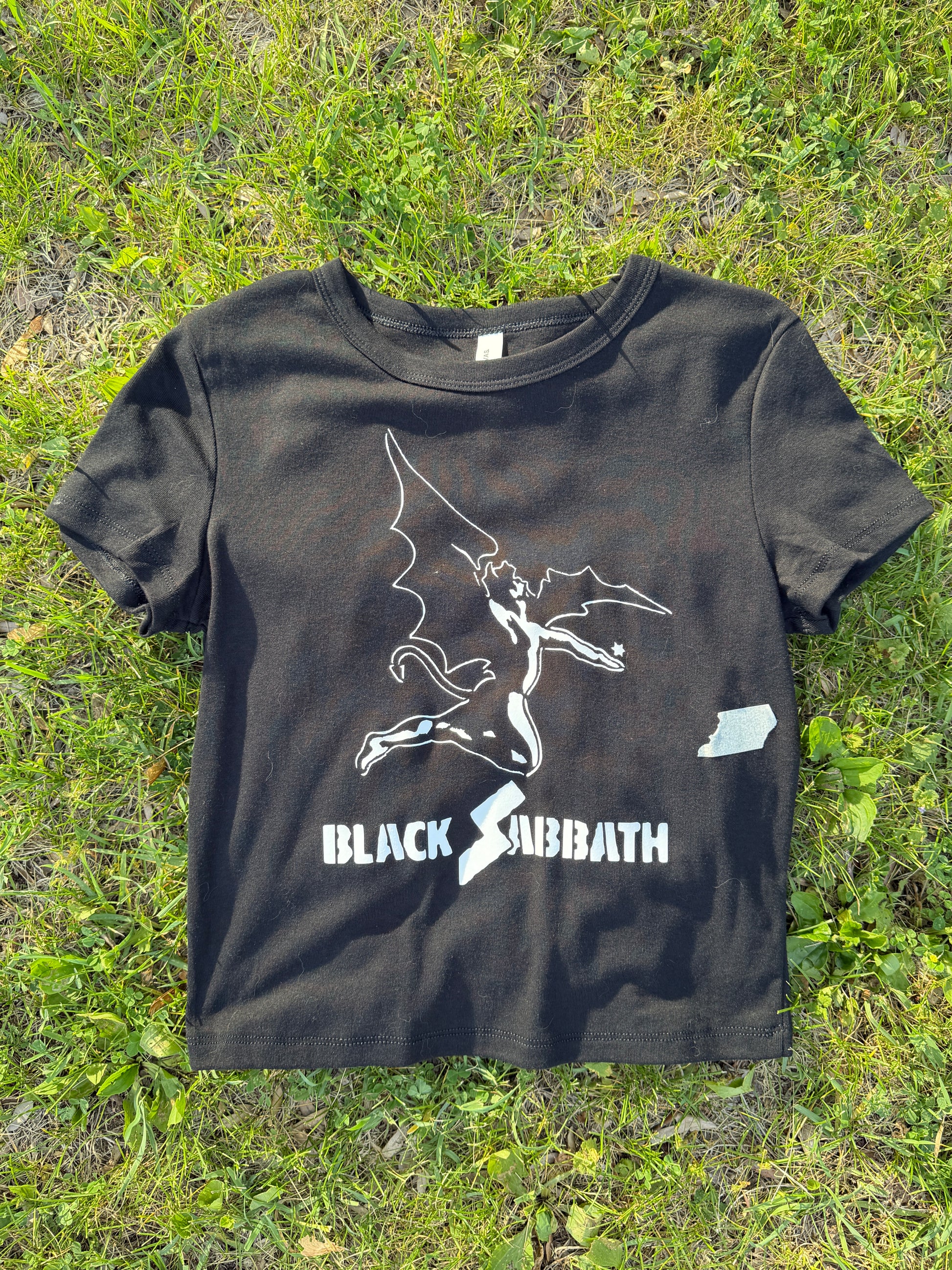A black t-shirt with a white graphic design of a bat and the text "BLACK SUBBATH" printed on it, laid out on a grassy surface.